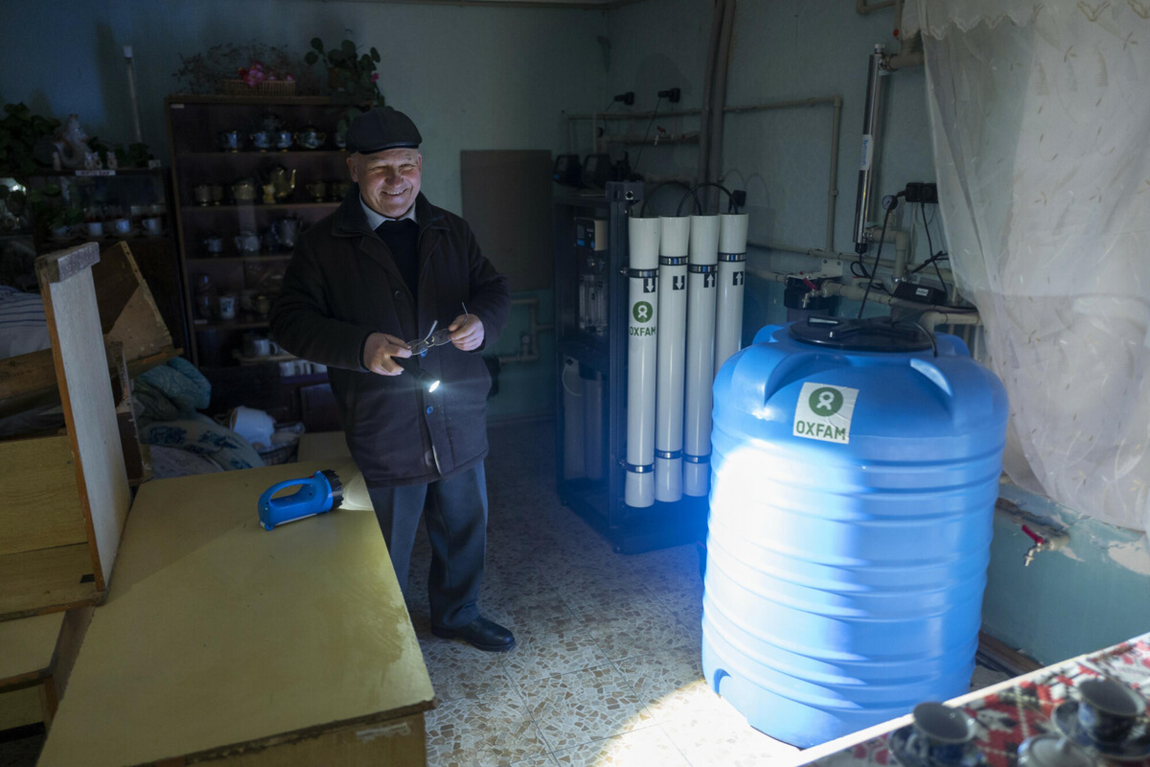 Volodymyr Yakovlev steht in einem dunklen Raum. mit einer Taschenlampe zeigt er eine Wasseraufbereitungsanlage auf der Oxfam steht. Er lacht zufrieden.