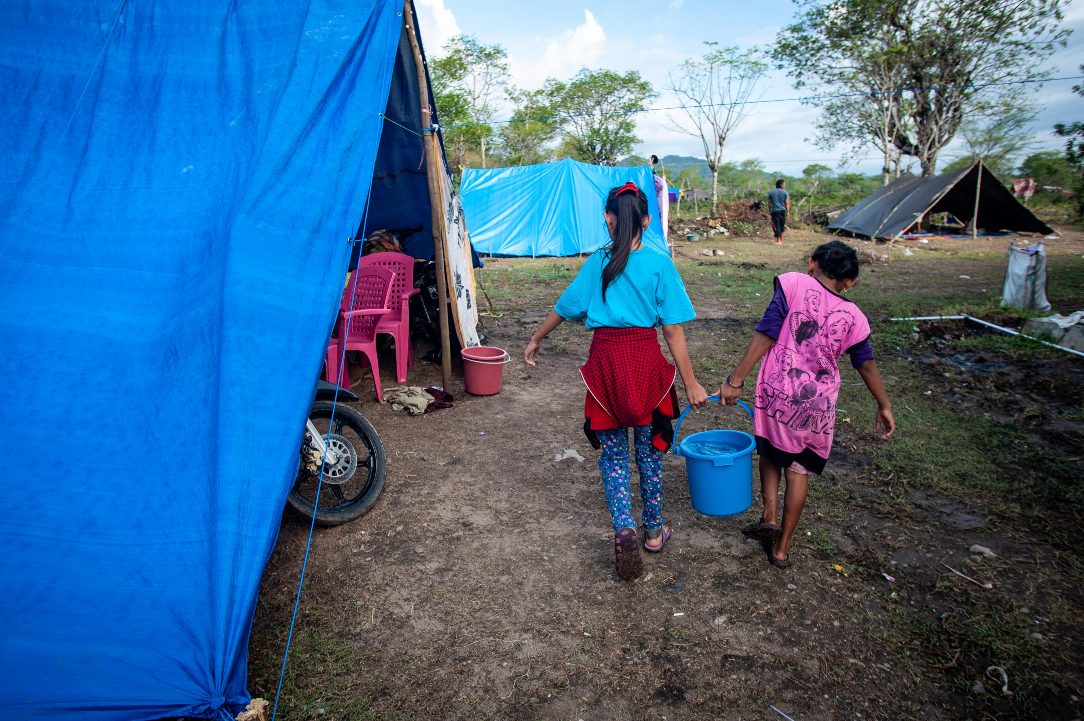 Zwei Mädchen tragen einen Eimer Wasser zwischen Zelten.