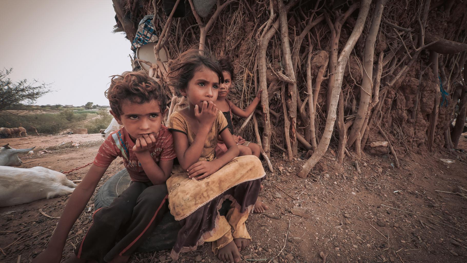 Drei Kinder sitzen vor einer einfachen Unterkunft aus Holz und abgenutzten Tüchern