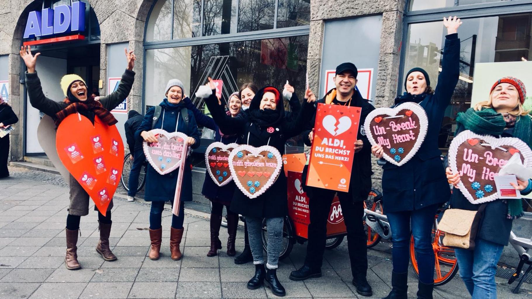 Oxfam-Aktivist*innen vor einer Aldi-Filiale halten Plakate und Lebkuchenherzen mit der Aufschrift „Aldi, du brichst uns das Herz“, „#aldigebrochenenherzen“ und „Un-Break My Heart“