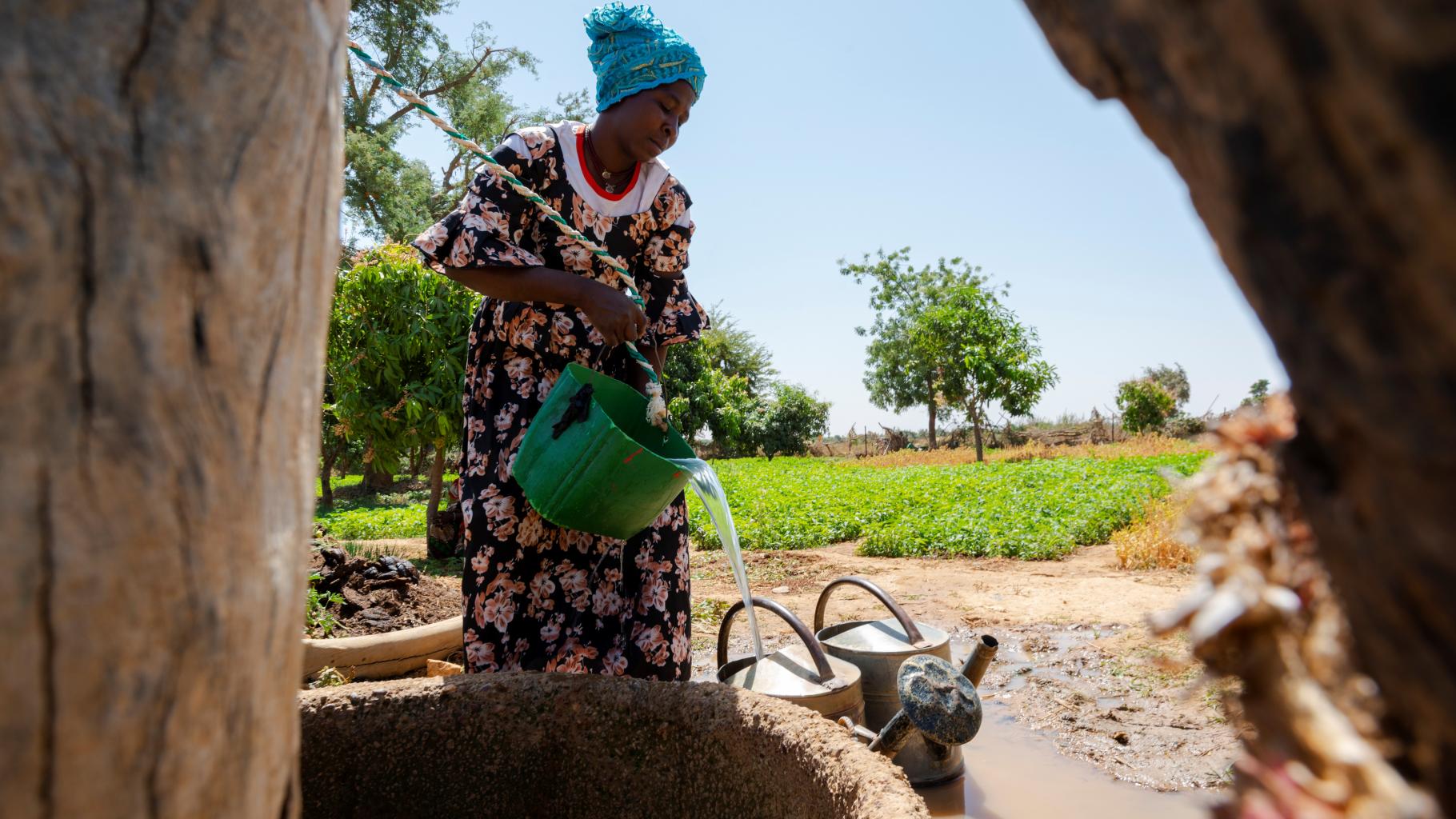 Aminiata Diallo befüllt Gießkannen mit Wasser. Im Hintergrund sind grüne Anbauflächen zu sehen.
