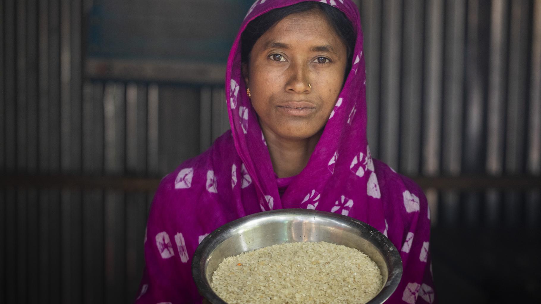 Eine Frau guckt in die Kamera und hält uns eine Schüssel mit Reiskörnern entgegen