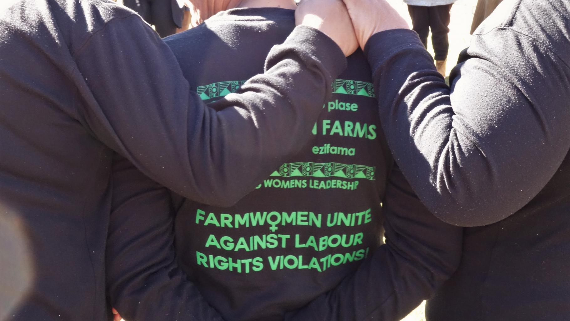 Drei Frauen stehen Arm in Arm mit dem Rücken zu uns, auf ihren T-Shirts steht "Farmwomen unite against labour rights violations" 