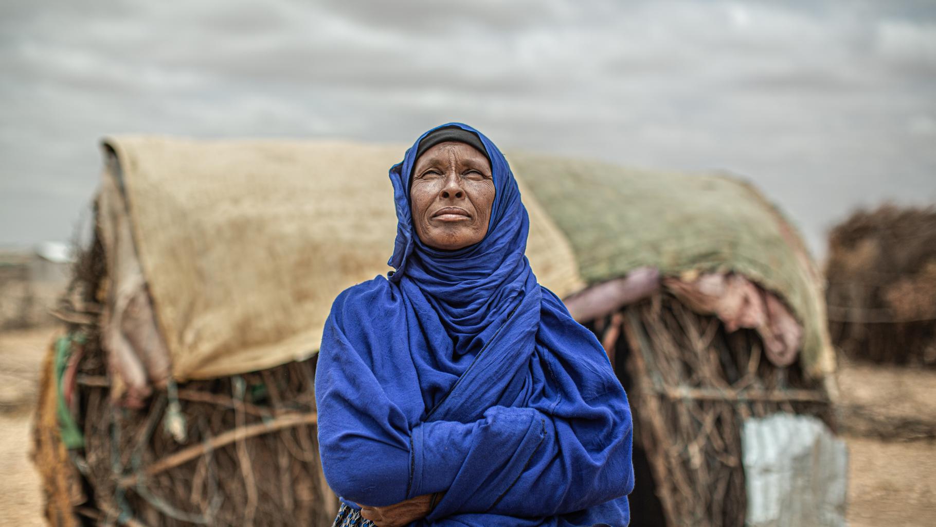 Amina Ibrahim schaut in einen wolkenbedeckten Himmel und verschränkt die Arme vor der Brust. Im Hintergrund ist ein einfaches Haus aus Decken und Holz zu sehen. 