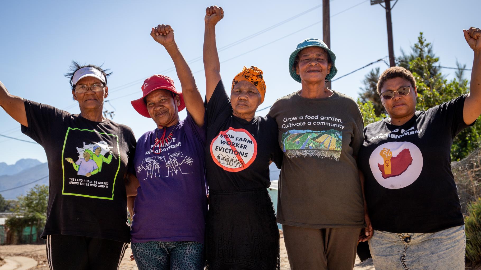 Gruppenbild von Women on Farms Project Aktivistinnen. Sie strecken ihre Fäuste in die Luft. In der Mitte ist Bettie Fortuin zu sehen, sie trägt ein gelbes, geknotetes Kopftuch und ein schwarzes T-Shirt mit der Aufschrift "Stop Farm Worker Evictions"