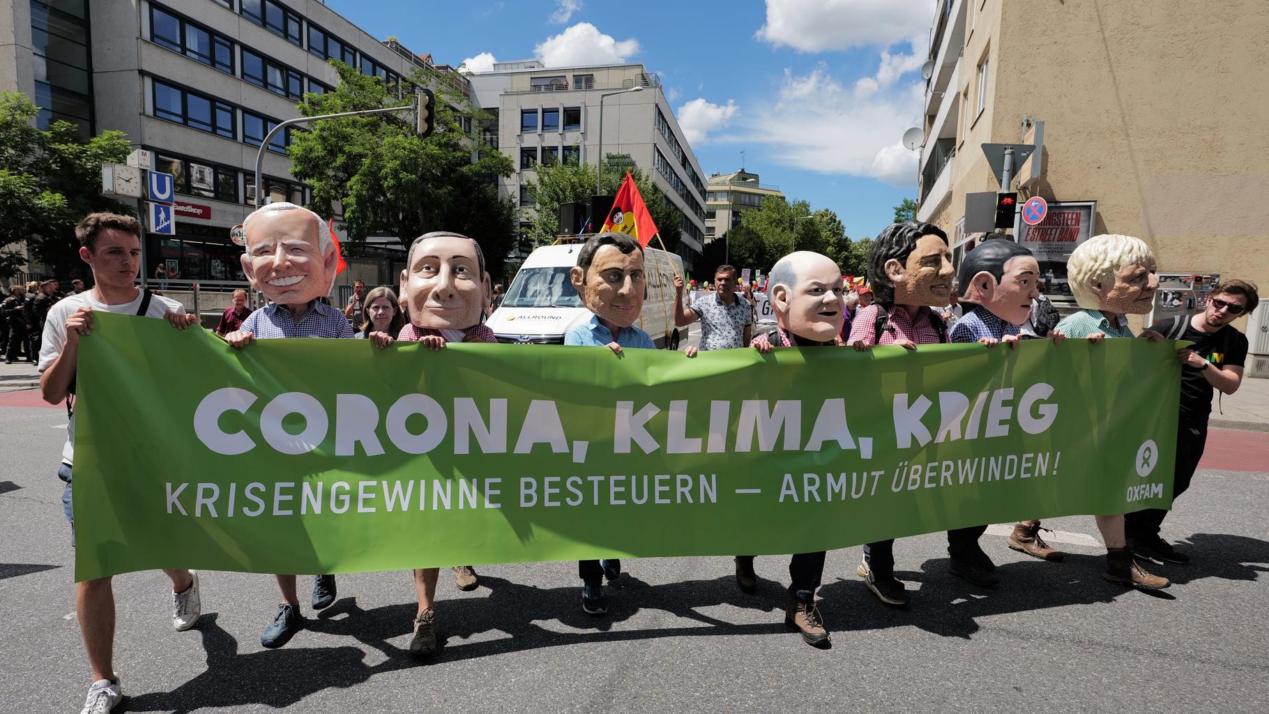 9 Oxfam-Mitarbeiter*innen tragen einen grünen Banner mit der Aufschrift "Corona, Klima, Krieg: Krisengewinne besteuern - Armut überwinden" auf der G7-Demo 2022. 7 von ihnen (mittig) tragen Big-Heads und posieren so als G7-Regierungschefs.