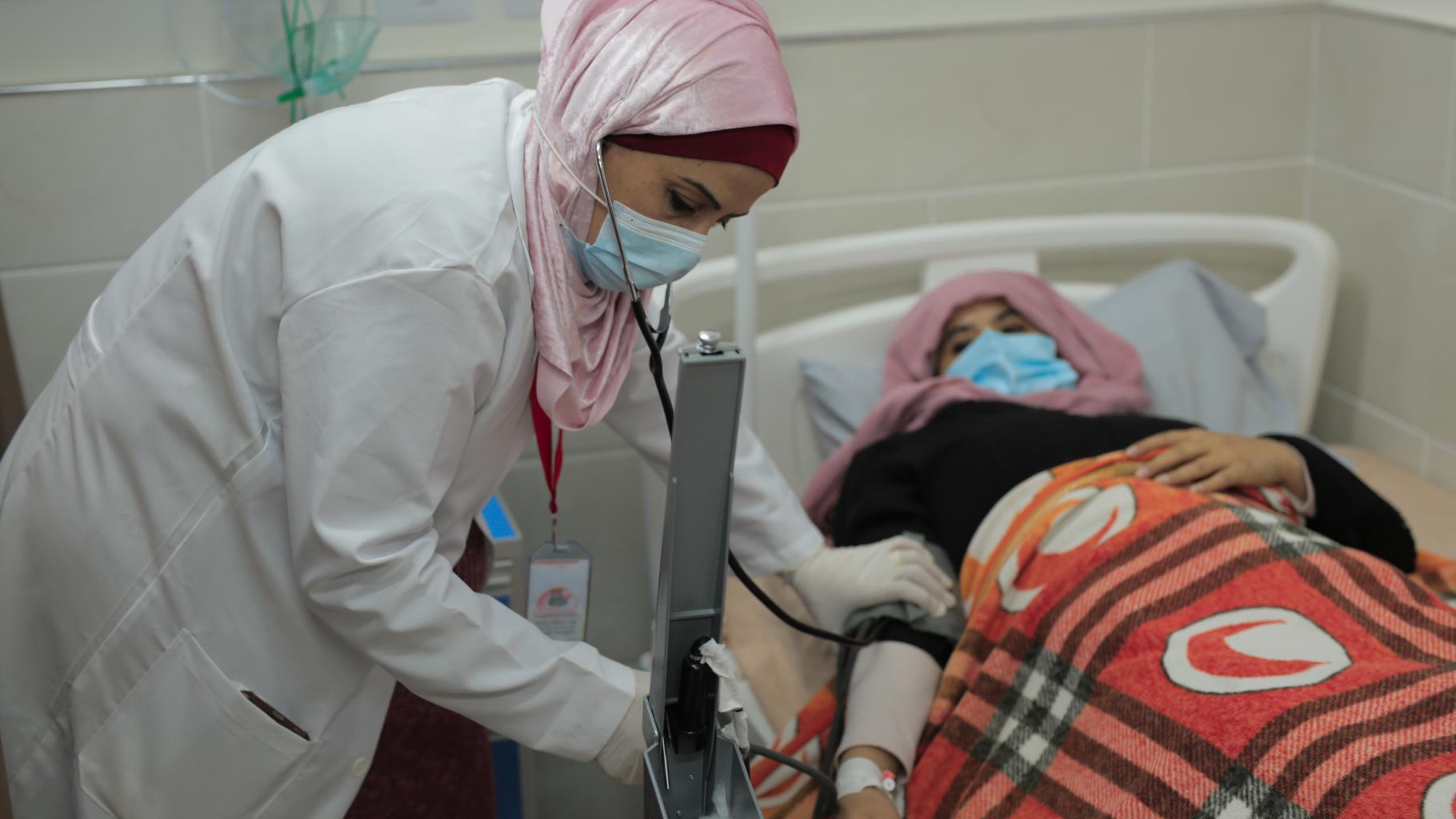 Heba Shalan, bekleidet mit einem weißen Kittel, misst Blutdruck einer Patientin, die in einem Krankenhausbett liegt. Beide tragen einen Mundschutz.
