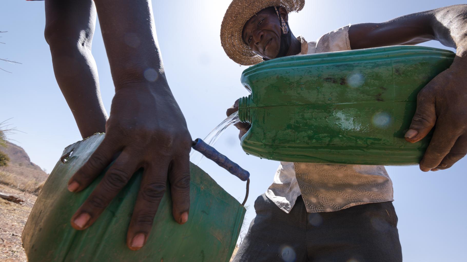 Samuel Sawadogo teilt Wasser auf zwei grüne Wasserkanister auf. Er trägt einen Hut zum Schutz vor der Sonne, ein beige-gemustertes Hemd und eine dunkle Hose. Man sieht links die Hände einer weiteren Person, die den zweiten Kanister hält.