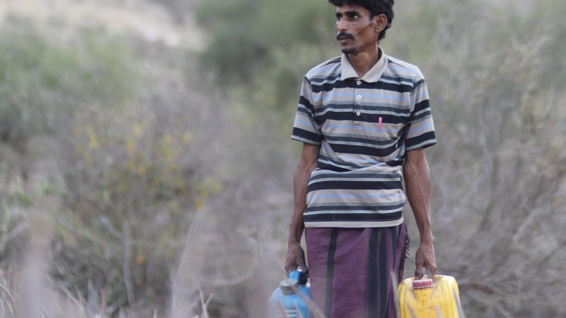 Ali trägt zwei Wasserkanister durch eine trockene Landschaft.
