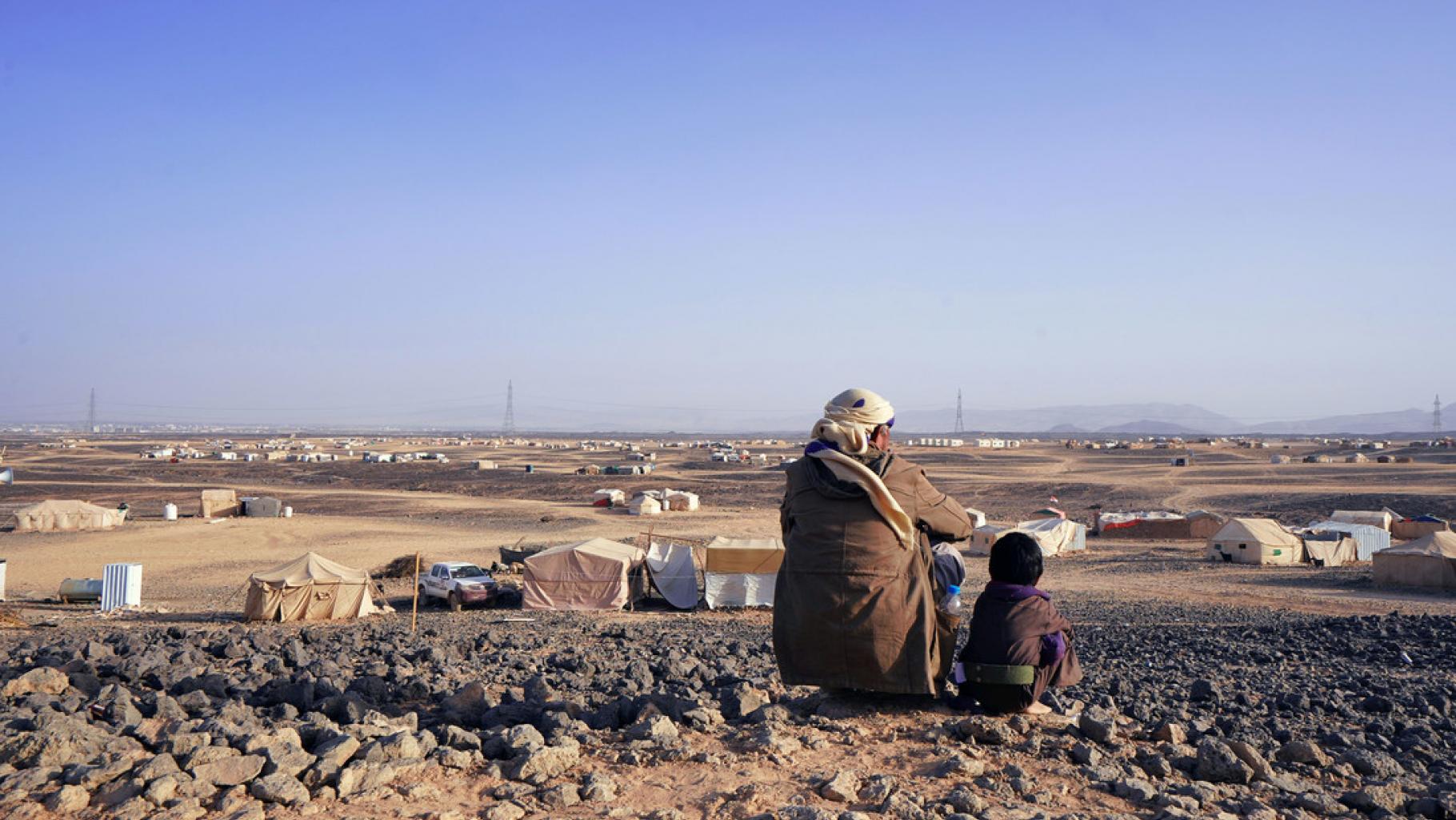 Ein Vater und sein siebenjähriger Sohn sitzen auf einem Hügel und schauen hinunter auf das Geflüchtetencamp Alswidan in Marib (Jemen). Weit und breit sind nur trostlose Steinwüste und einige Zelte zu sehen.