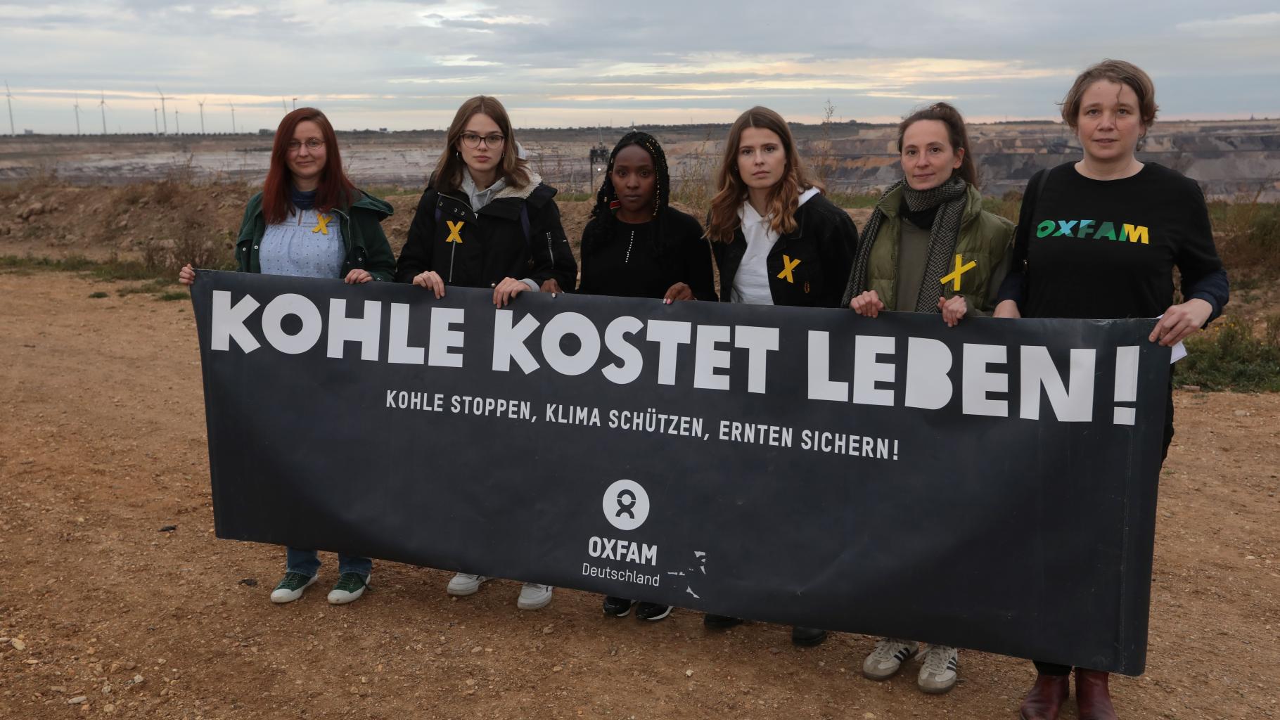 Sechs Frauen stehen mit einem schwarzen Banner in der Hand am Rand des Tagebaus Garzweiler. Auf dem Banner steht: Kohle kostet leben! Kohle stoppen, Klima schützen, Ernten sichern!