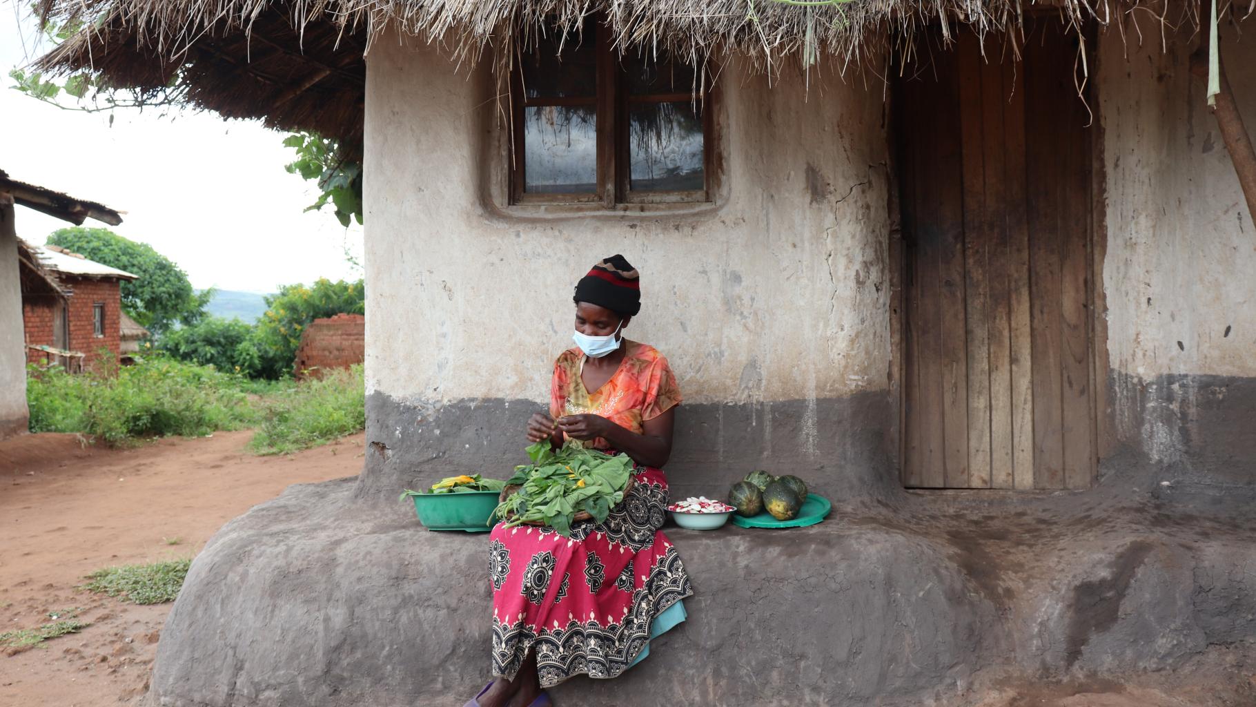 Loveness N'Dindo. mit einem rot-schwarz gemusterten Rock und orangenen T-Shirt sowie Mundschutz bekleidet, sitzt auf einer Steinbank vor ihrem Haus. Auf ihrem Schoß sortiert sie Gemüse. Neben ihr befinden sich weitere Schüsseln mit Früchten. 