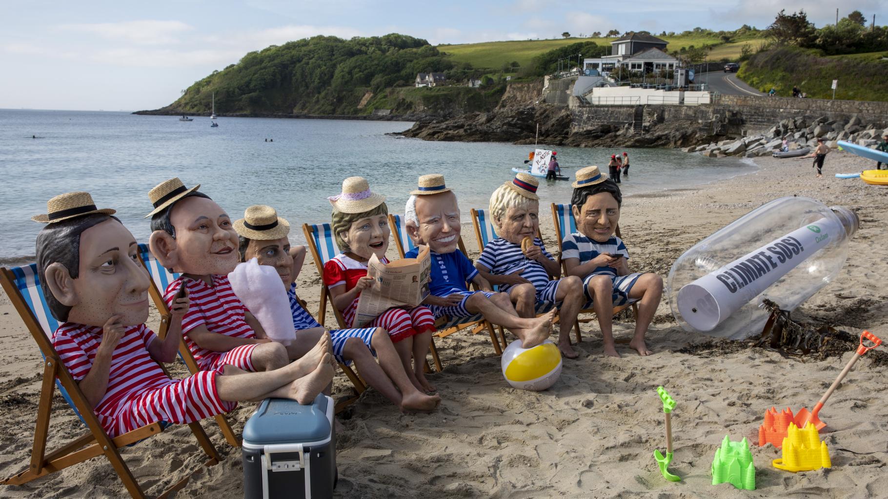Sieben Aktivist*innen sitzen in Liegestühlen an einem Strand.Sie tragen Badeanzüge und große Masken mit den Gesichtern der G/-Staats- und Regierungschef*innen.Neben ihnen liegt eine große Flaschenpost mit der Aufschrift „Climate SOS!“