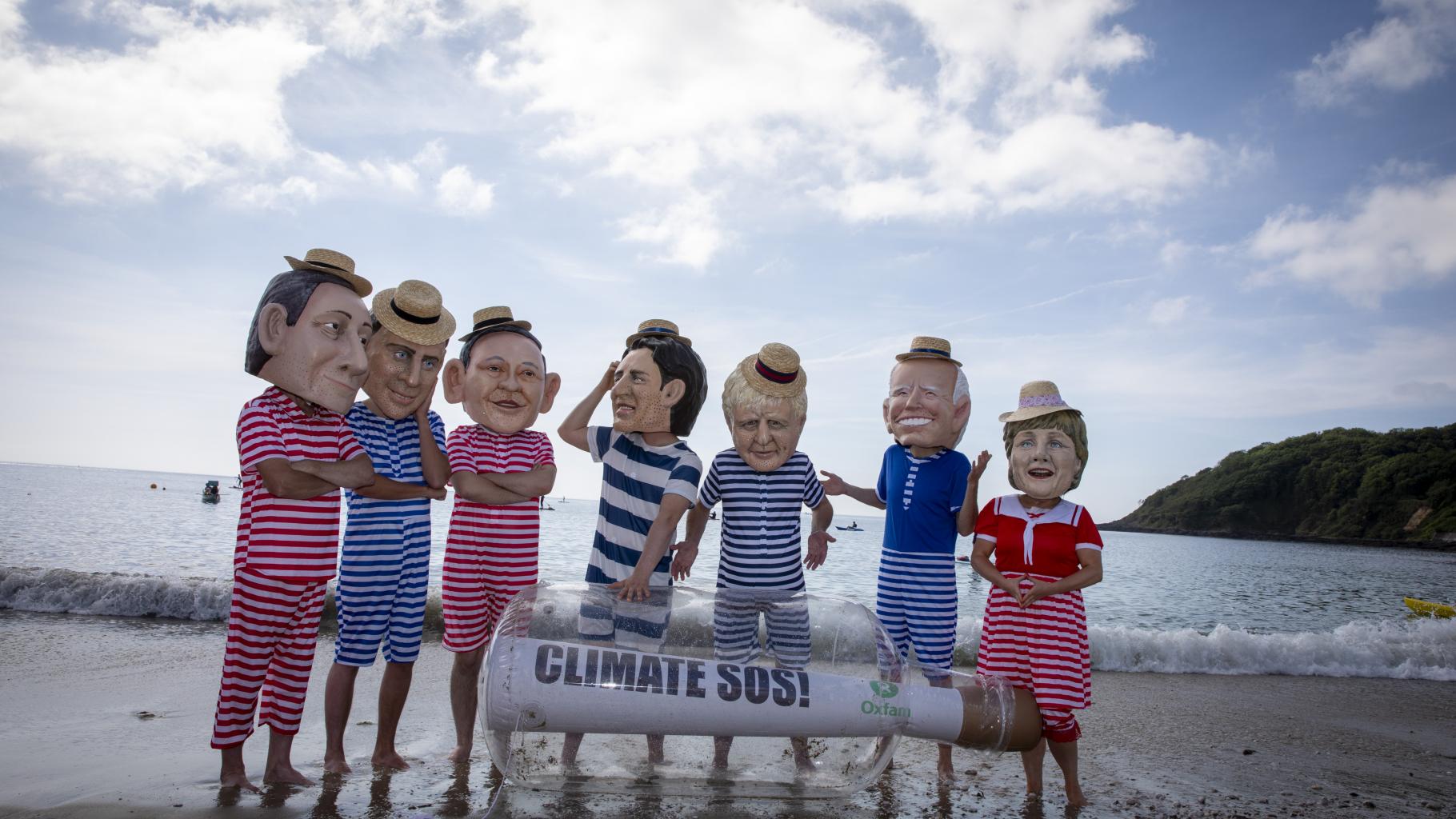 Aktivist*innen posieren als G7-Regierungschef*innen am Strand. In gestreiften Badesachen und Sonnenhüten stehen sie um eine riesige Flaschenpost herum und schauen sich fragend oder skeptisch an.