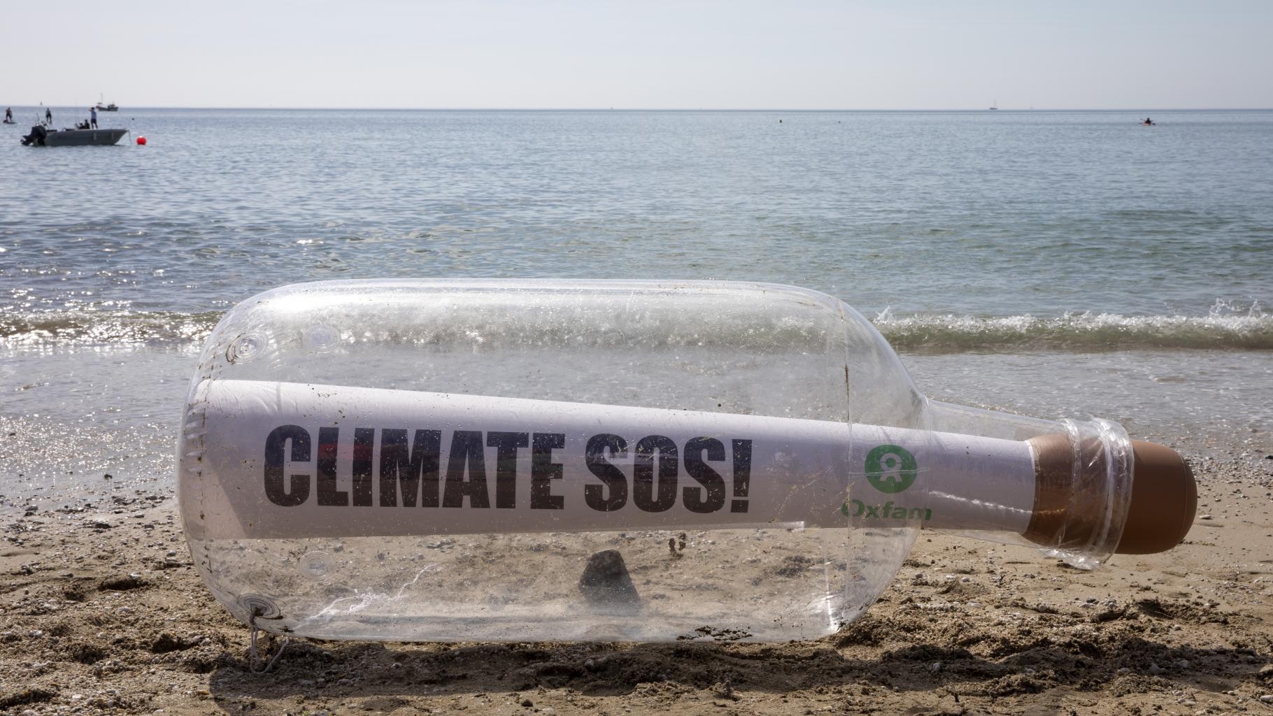 Eine riesige Flaschenpost mit der Aufschrift "Climate SOS" liegt an einem Strand. Im Hintergrund ist das Meer zu sehen. 