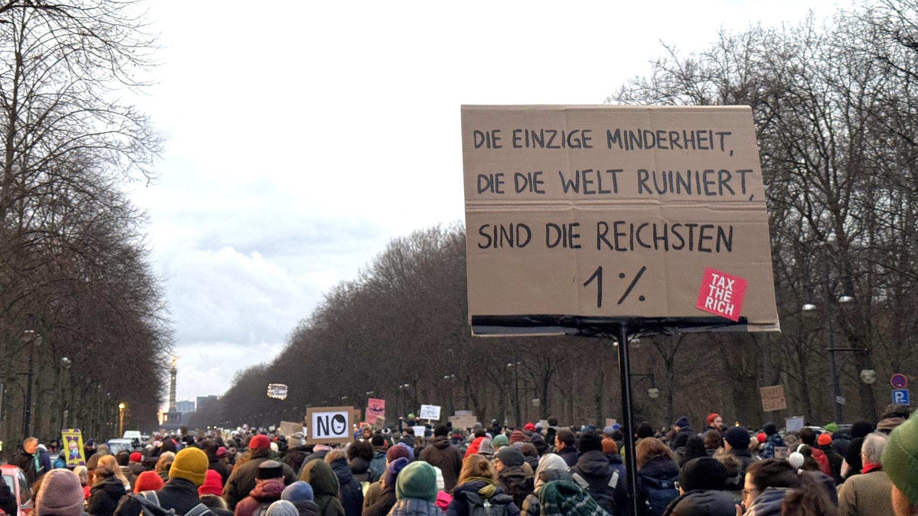 viele Menschen mit Demoschildern bewegen sich auf den Großen Stern in Berlin zu. Auf einem Schild steht "Die einzige Minderheit, die die Welt ruiniert, sind die reichsten 1 %"