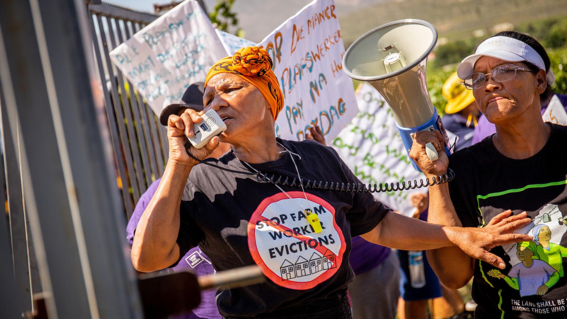 Eine Frau spricht in ein Megafon, die andere hält das Megafon. Aufschrift auf einem T-Shirt: „Stop Farm Worker Evictions“ „(Stoppt die Zwangsräumung von Farmarbeiter*innen“)