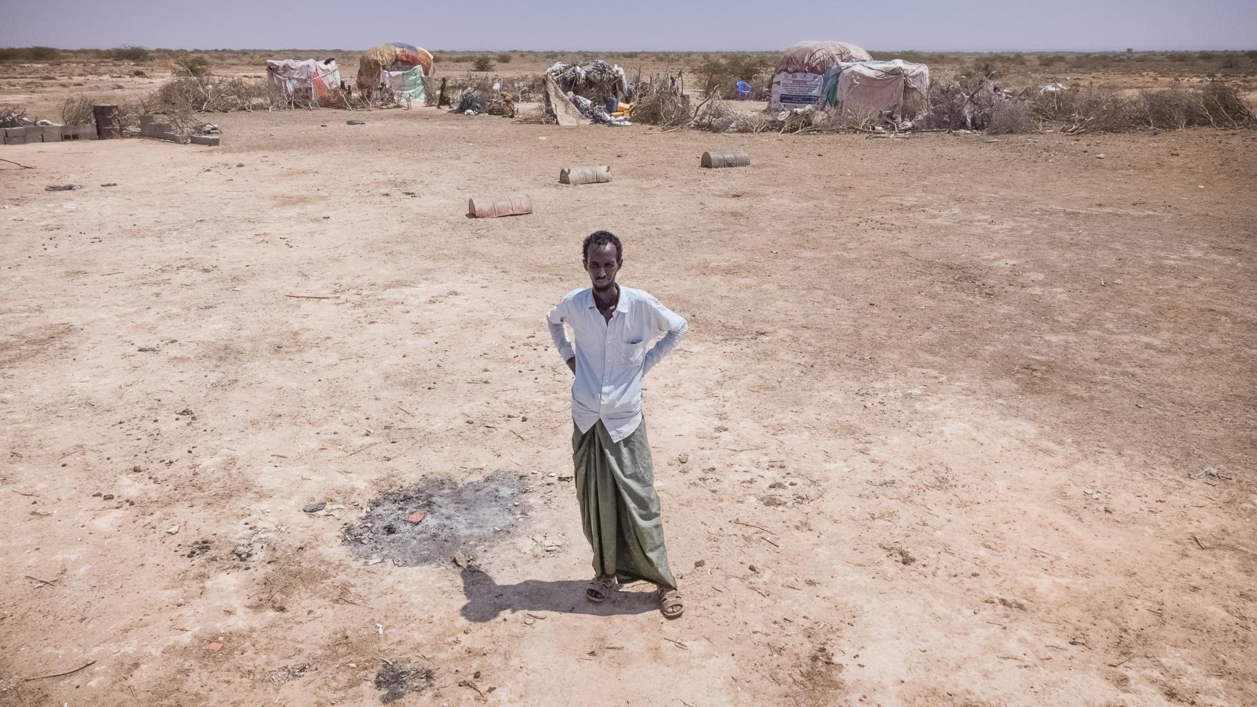 Abdulahi Farah Isse steht, bekleidet mit einem weißen Shirt und einem grauen Wickelrock, auf einem sehr trockenen Boden. Im Hintergrund ist seine Unterkunft zu sehen.