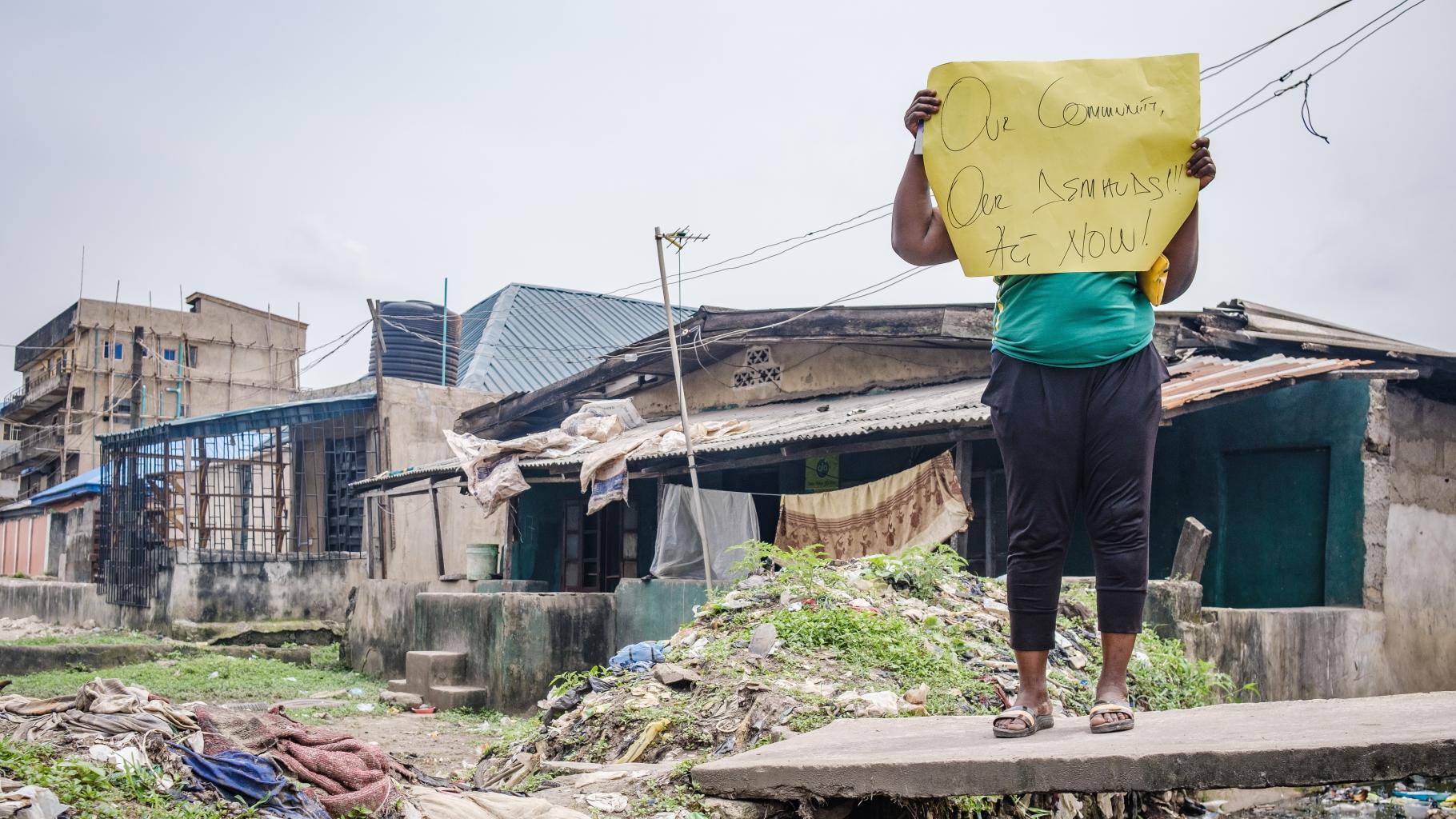 Eine Aktivistin hält ein Plakat mit der Aufschrift "Unsere Community, unsere Forderungen! Jetzt handeln!" hoch. Sie trägt ein grünes T-Shirt und eine dunkle Hose. Im Hintergrund sind Häuser zu sehen.