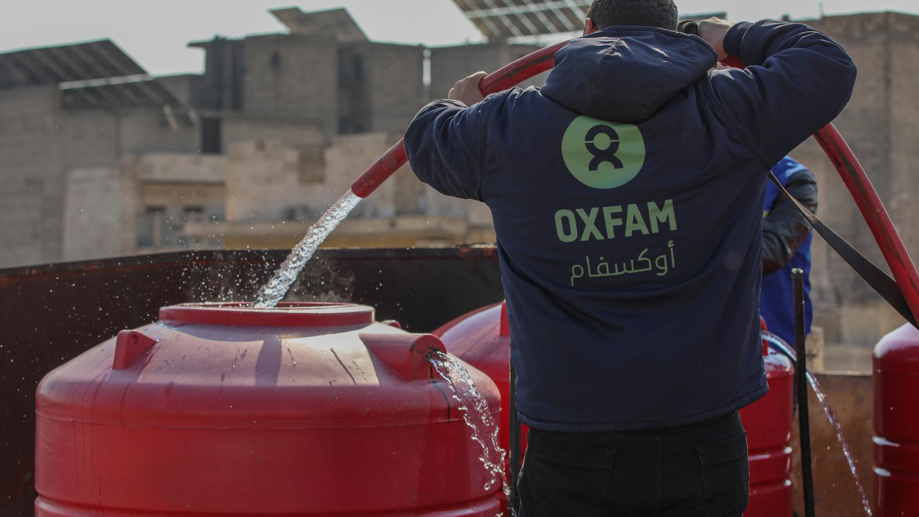 Ein_e Oxfam-Mitarbeiter_in, mit einer blauen Kaputzenjacke mit grünem Oxfam-Logo bekleidet, befüllt mithilfe eines Schlauches einen roten Wassertank mit sauberem Trinkwasser.