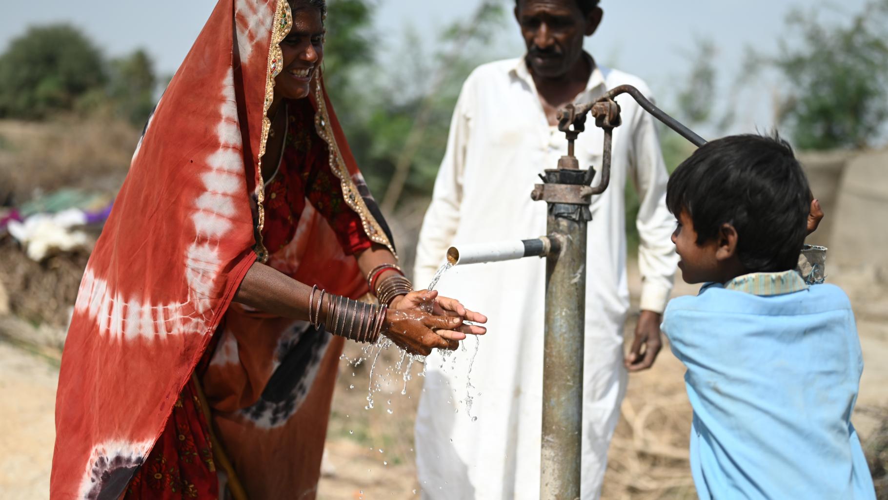 Eine Familie benutzt eine neu installierte Hand-Wasserpumpe. Das Kind (rechts) bedient die Pumpe, während die Mutter (links) ihre Hände unter den Wasserstrahl hält. Im Hintergrund steht eine dritte Person.