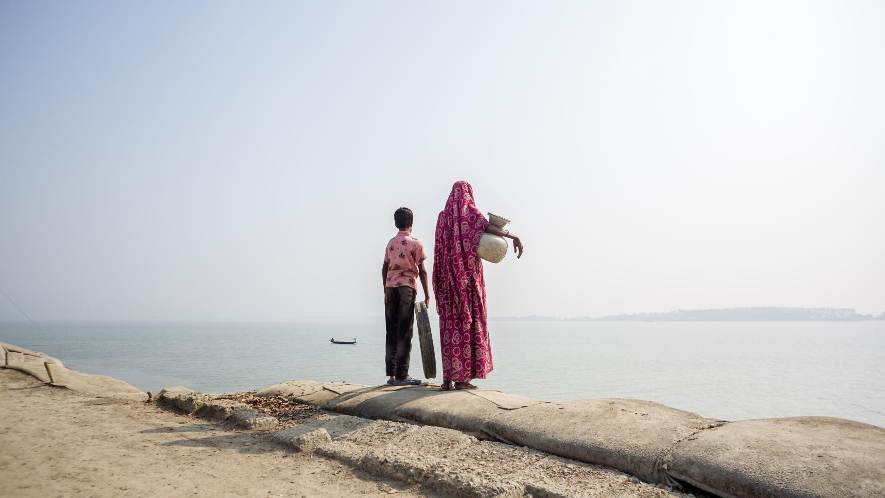 Eine Mutter steht mit ihrem Kind am Rande einer Küste. Unter dem Arm trägt sie einen Krug zum tranportieren von Wasser.