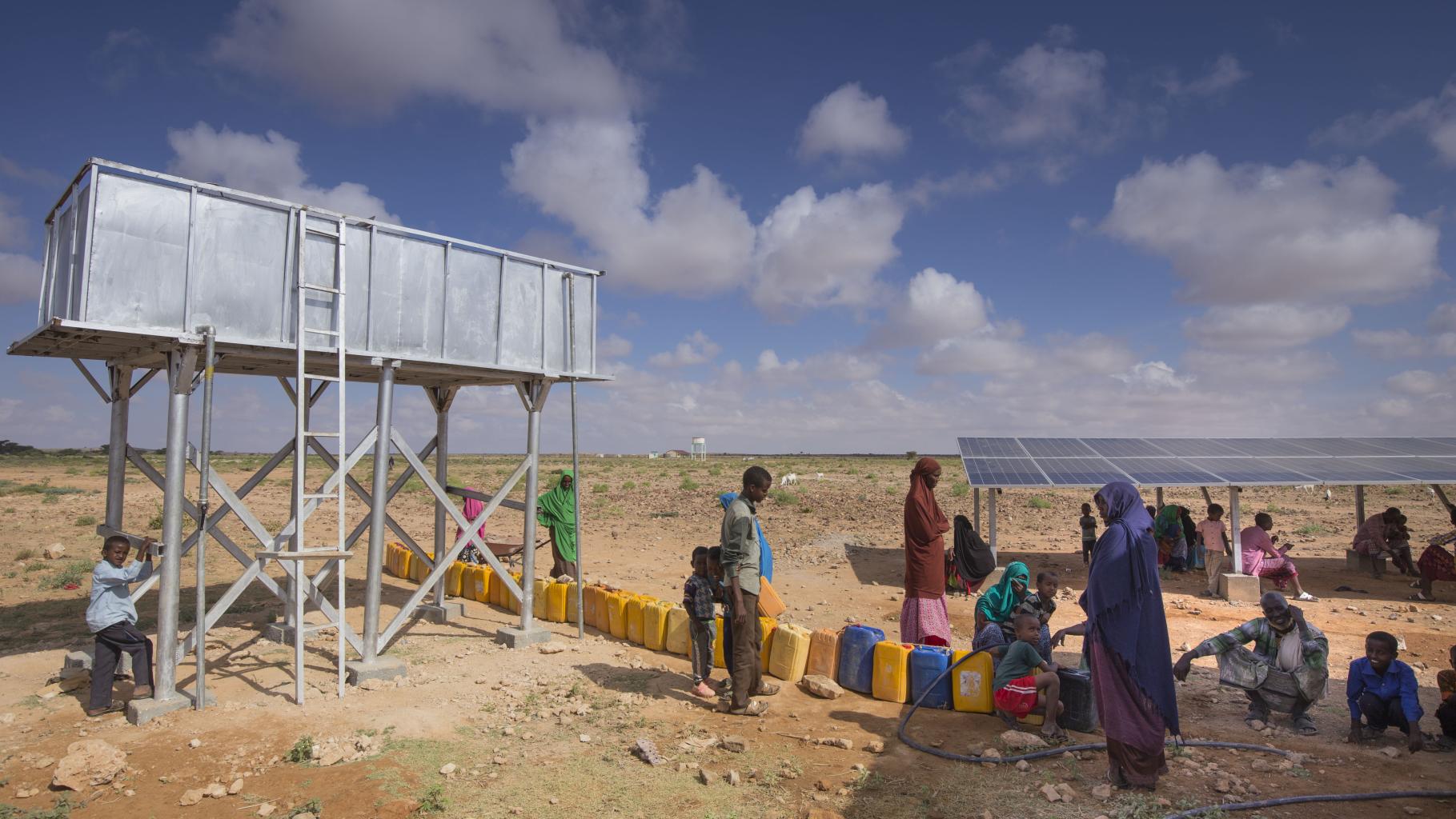 In der von Oxfam gebauten Entsalzungsanlage in Ceel Midgaan, Somaliland, wird sauberes Wasser erzeugt.Das Bild zeigt einen Wassertank, vor dem viele Wasserkanister stehen. Im Hintergrund steht eine Solaranlage, die die Entsalzungsanlage mit Strom versorgt. Unter den Solarpanels sitzen einige Menschen im Schatten. Weitere Menschen stehen und Hocken in der Sonne und warten darauf, ihre Kanister befüllen zu können.