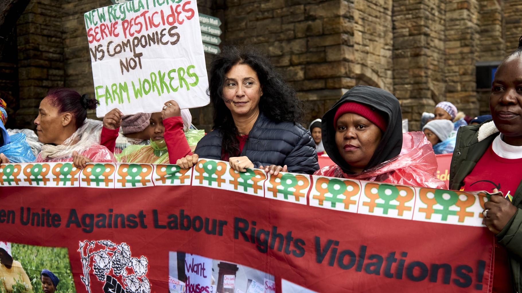 Frauen auf einer Demonstration, zu lesen ist „Unite against labor rights violations“ und „New regulations serve pesticides vs companies not farm workers“