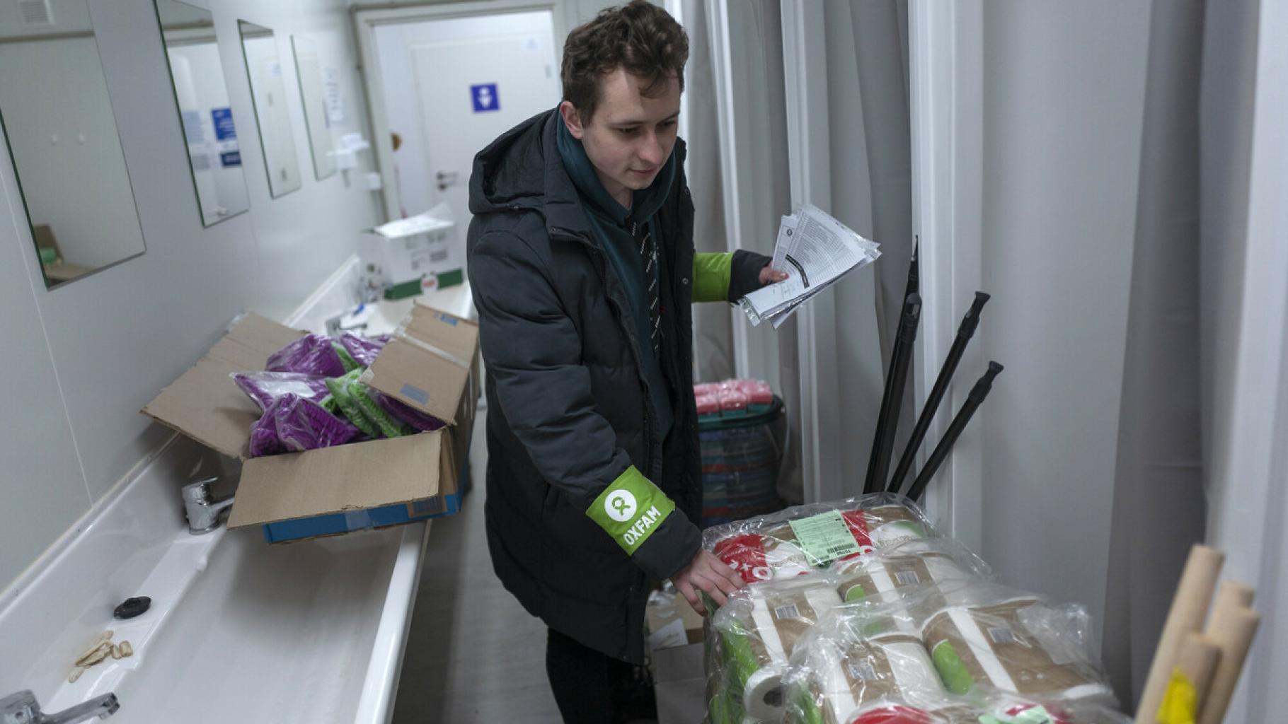 Vladislav Kubrak steht in einem Waschraum vor einem großen Paket Hygienepapierrollen. Hinter ihm befinden sich weitere Kartons mit Hygieneartikeln.