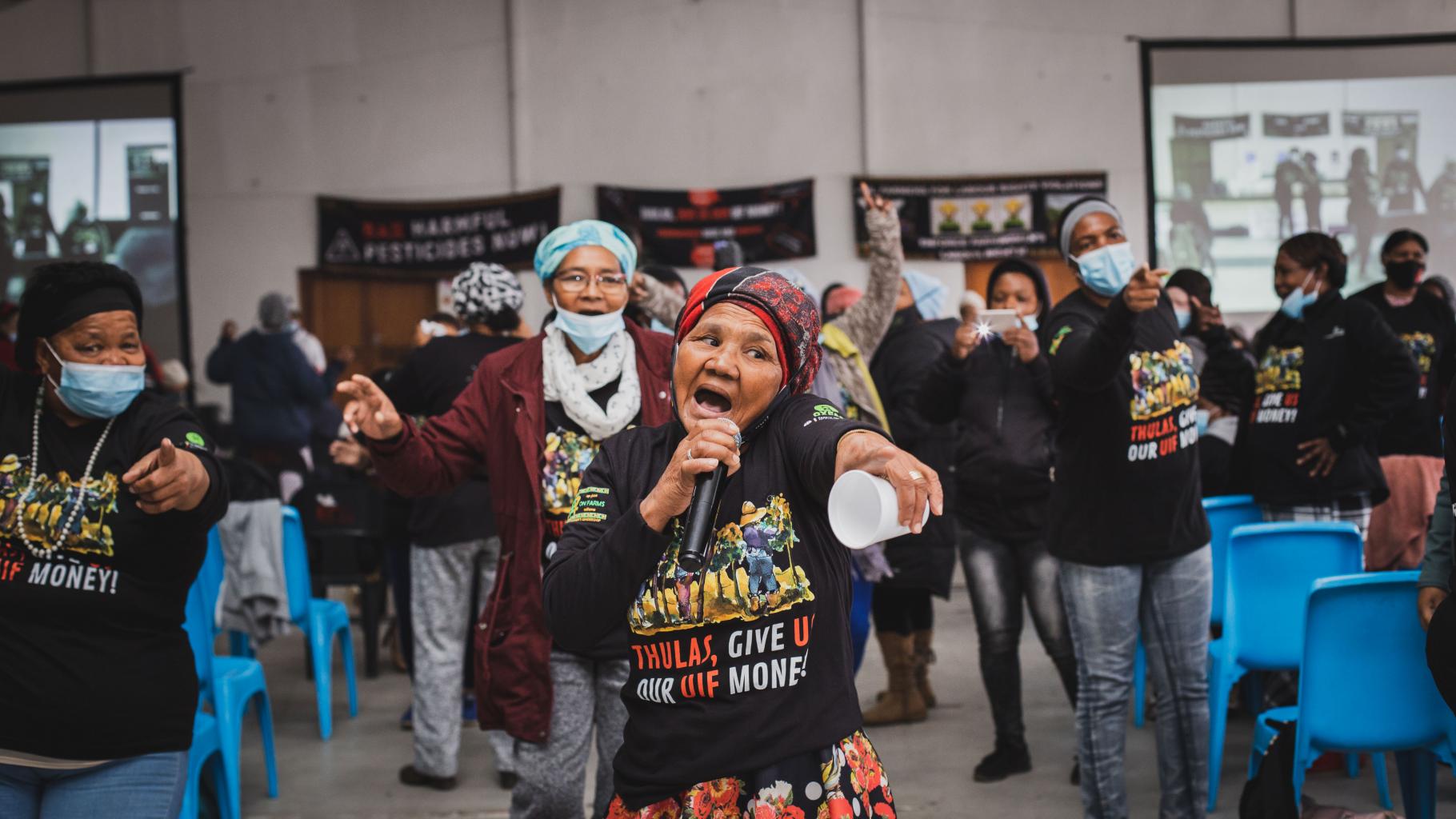 Eine Gruppe von Farmarbeiterinnen singen auf einer Protestaktion mit schwarzen Tshirts mit der Aufschrift "Thulas, give us our money".