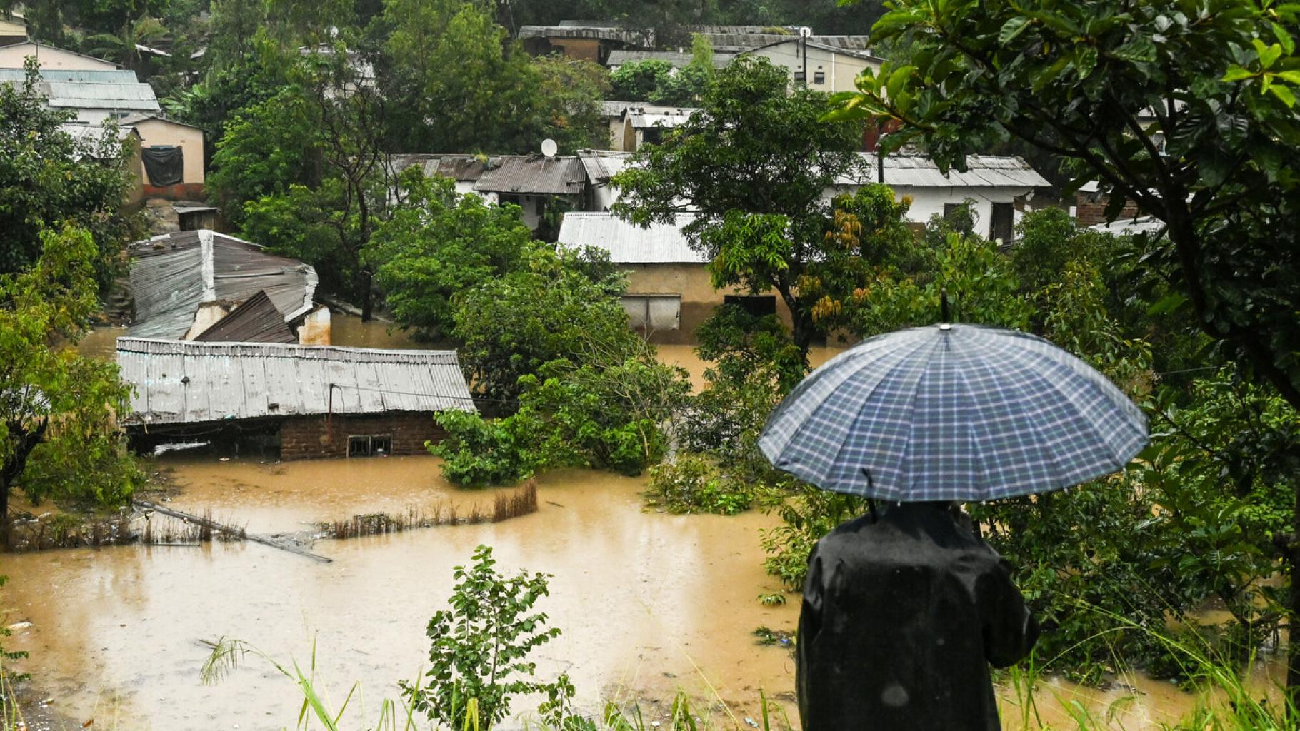 Eine Person mit Regenschirm steht auf einer Anhöhe. Hinter ihr sieht man Häuser, die teilweise bis zum Dach im Wasser stehen.
