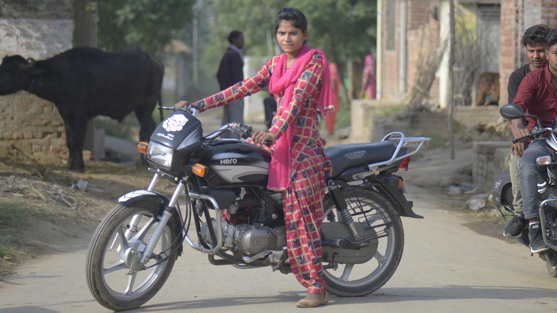 Eine junge indische Frau sitzt auf einem Motorrad; neben ihr zwei Männer auf einem anderen Motorrad