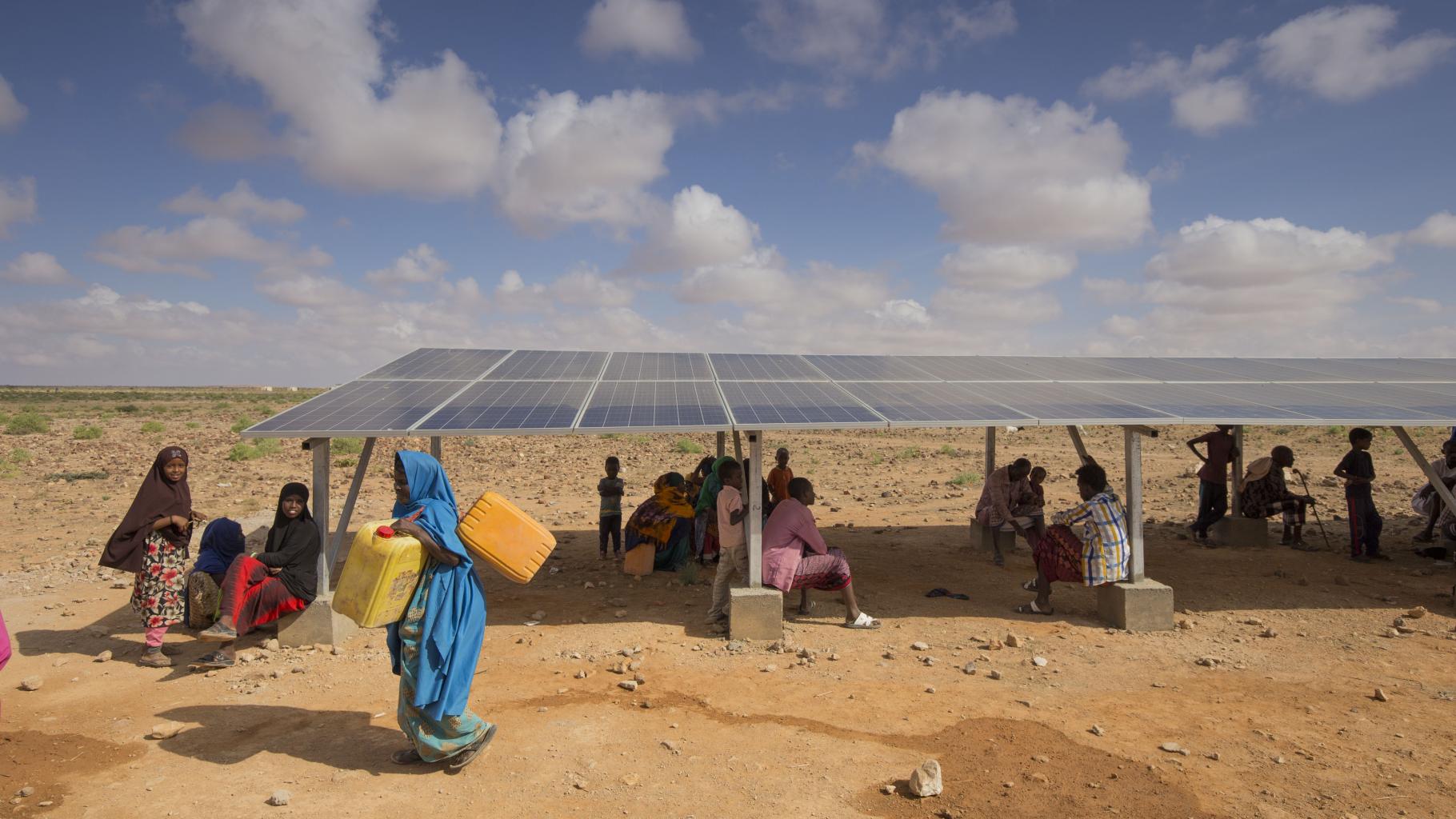 Ein Mädchen trägt Wasserkanister, während eine Gruppe von Menschen im Schatten einer Solaranlage sitzt