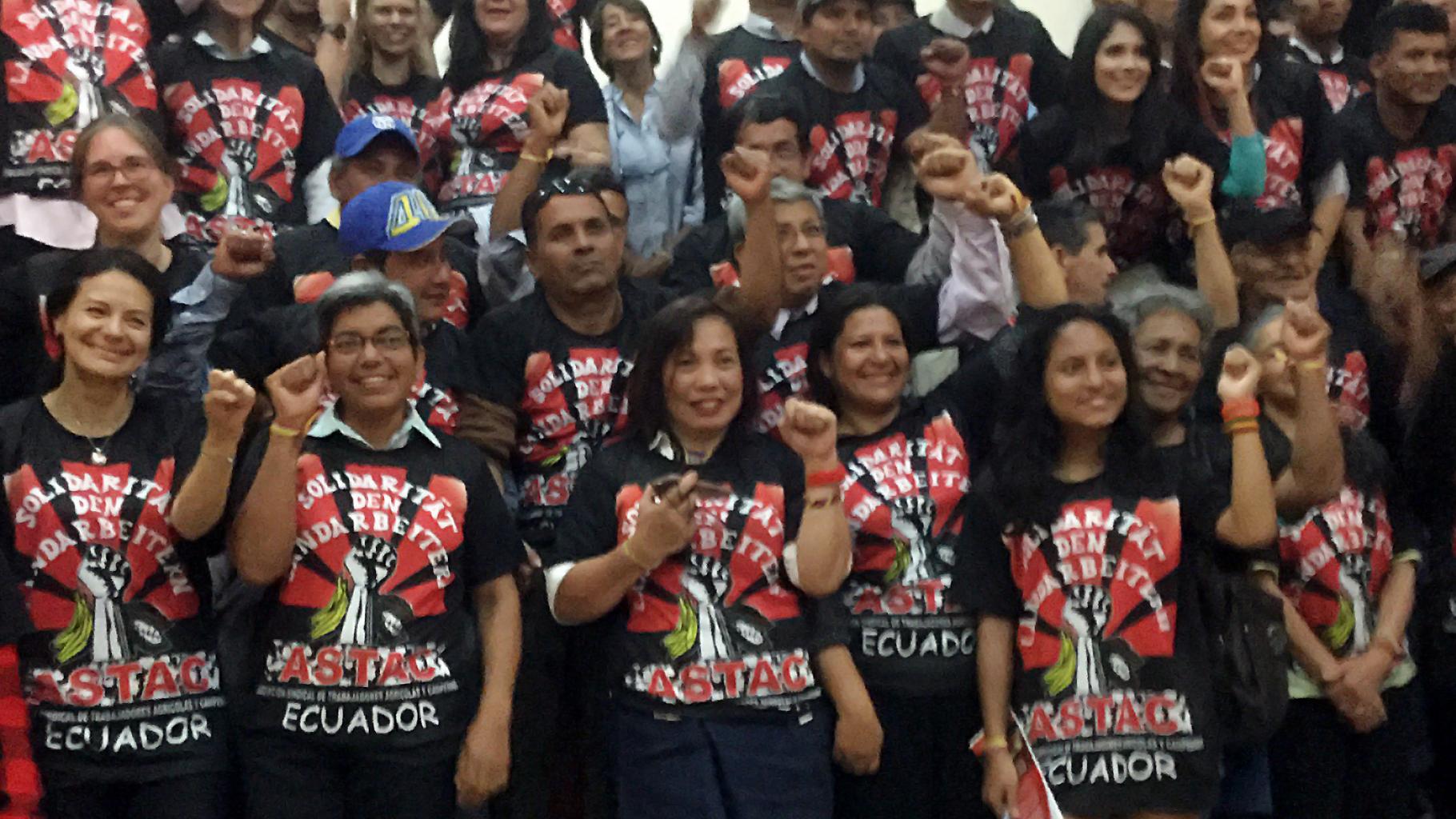 Gruppenfoto; alle tragen ein T-Shirt mit der Aufschrift „Solidarität mit den Landarbeitern – ASTAC – Ecuador“ und heben die Fäuste