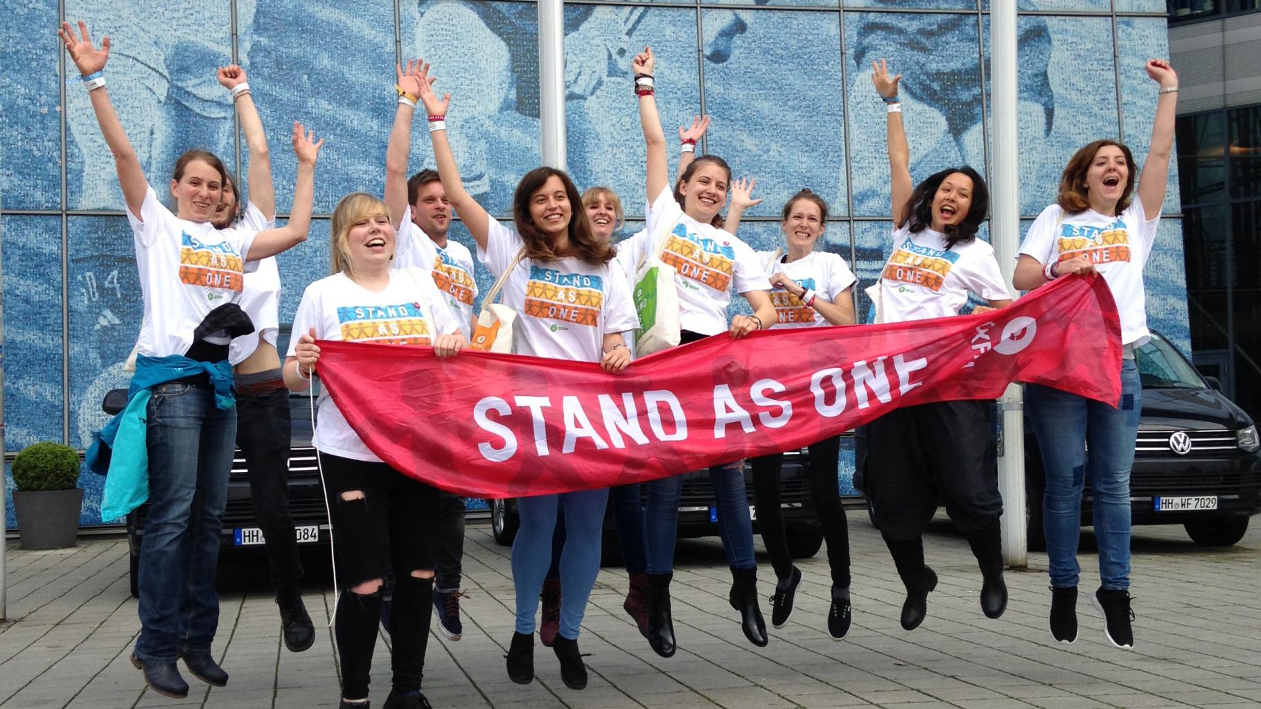Hüpfende Konzertaktivistinnen halten ein Banner mit der Aufschrift: Stand As One