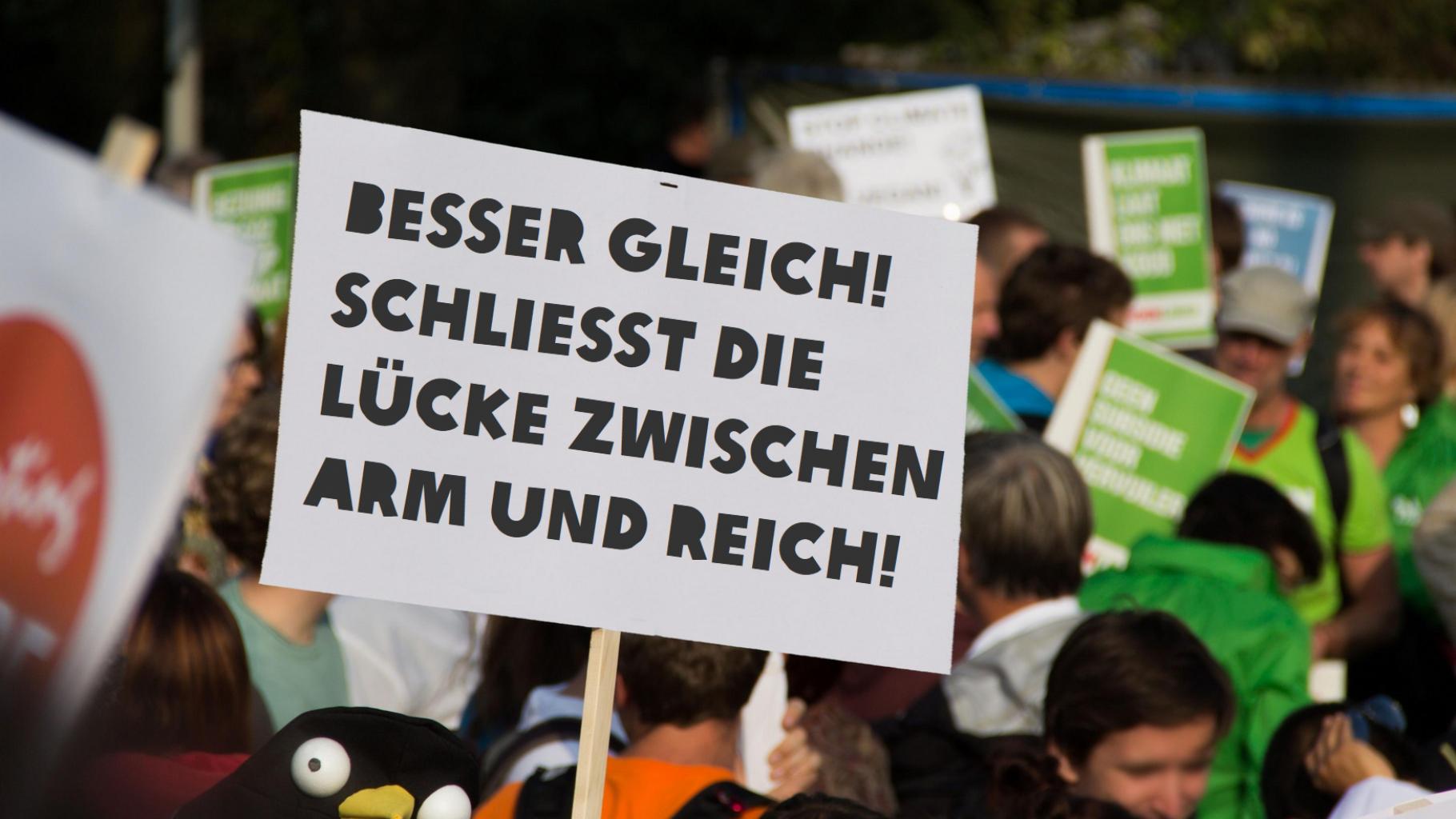 Im Vordergrund ein Schild mit der Aufschrift: „Besser Gleich! Schließt die Lücke zwischen Arm und Reich!“ Im Hintergrund Menschen auf einer Demonstration