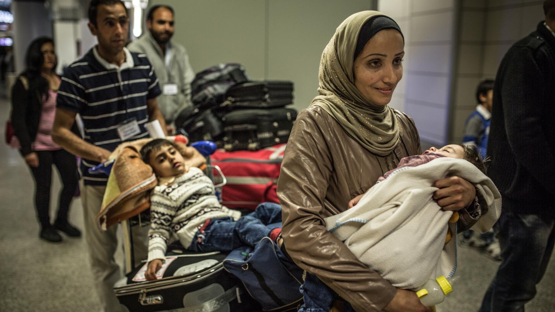 Eine Frau mit einem kleinen Kind auf dem Arm geht durch die Ankunftshalle eines Flughafens; hinter ihr schiebt ein Mann einen Kofferkuli mit Gepäck, auf dem ein müder Junge sitzt.