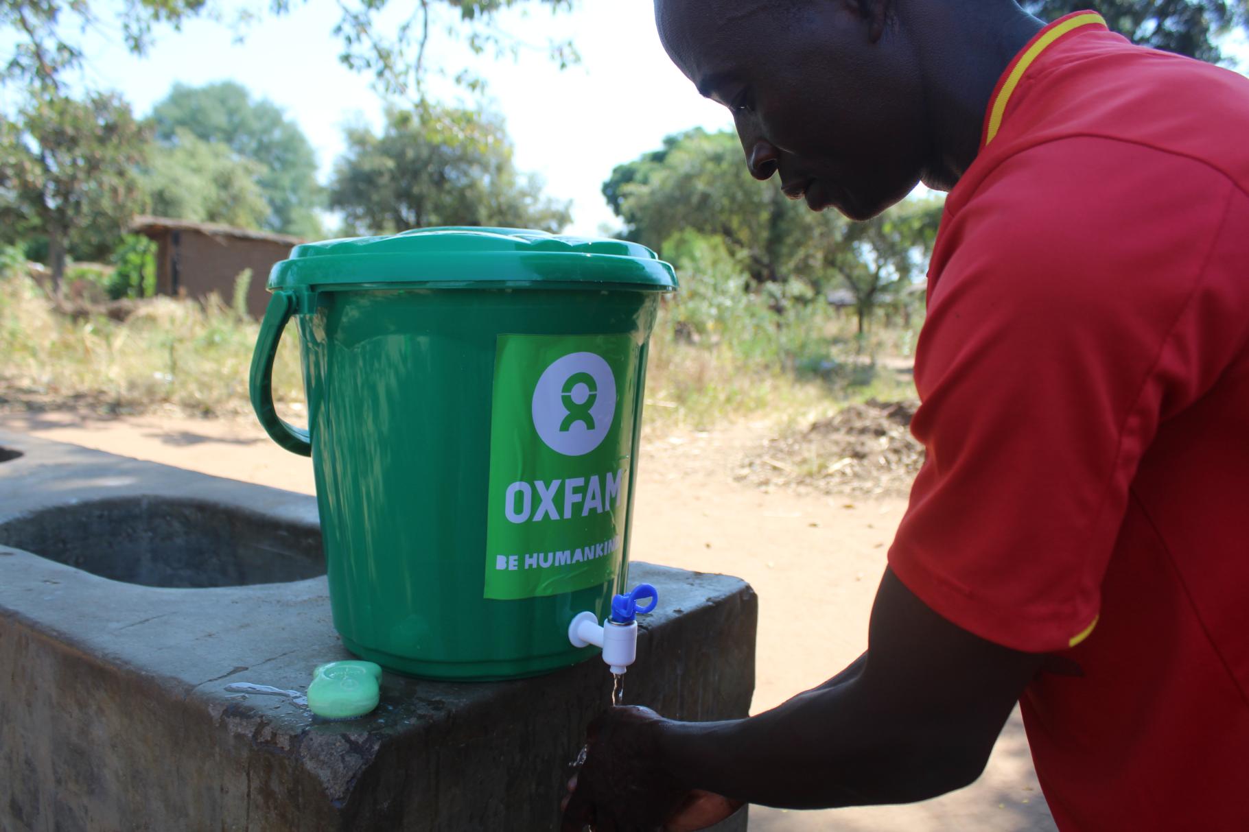 Rexo Chimotokoma aus Malawi demonstriert die Bedienung eines grünen Wassereimers mit Oxfam-Logo drauf.