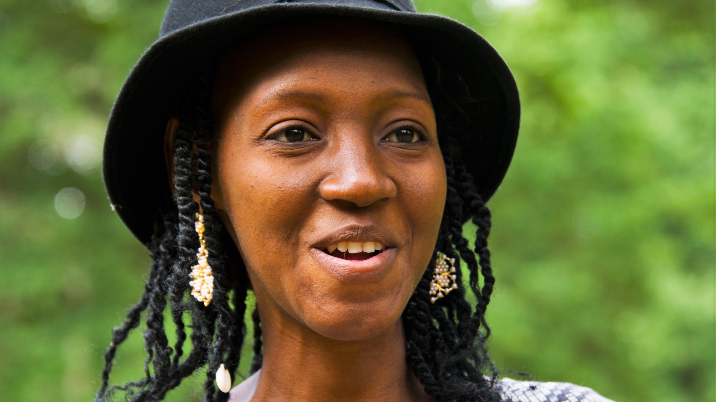 Porträt von Gogo Bambera. Sie trägt einen schwarzen Hut und ein grau-weißes Oberteil. Sie lächelt.