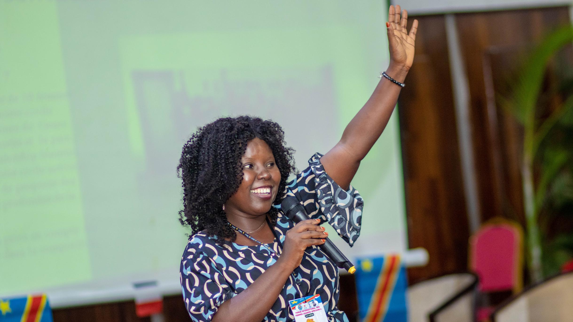Grâce spricht auf dem Internationalen Afrikanischen Frauenforum (FIFAF) in Kinshasa (Demokratische Republik Kongo). Sie hält ein Mikrofon in der rechten Hand, mit der Linken winkt sie Zuhörenden zu und lächelt sie an. Sie trägt eine blau-gemusterte Tunika.