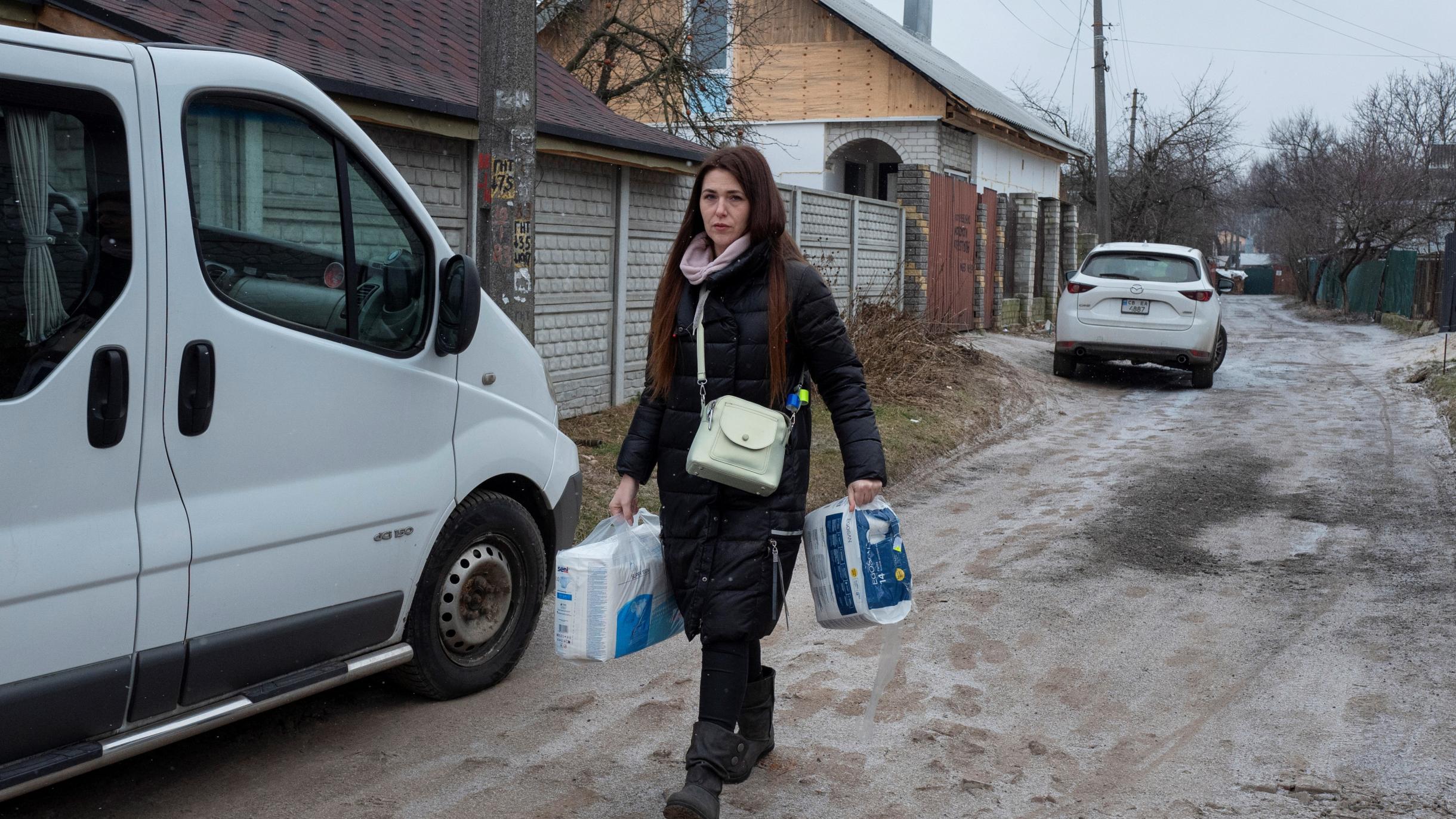 Ana Kulyeva läuft in Winterkleidung und mit Drogerieartikeln in der Hand eine Straße in Chernihiv entlang. Im Hintergrund sind zwei Autos und Häuser zu sehen.