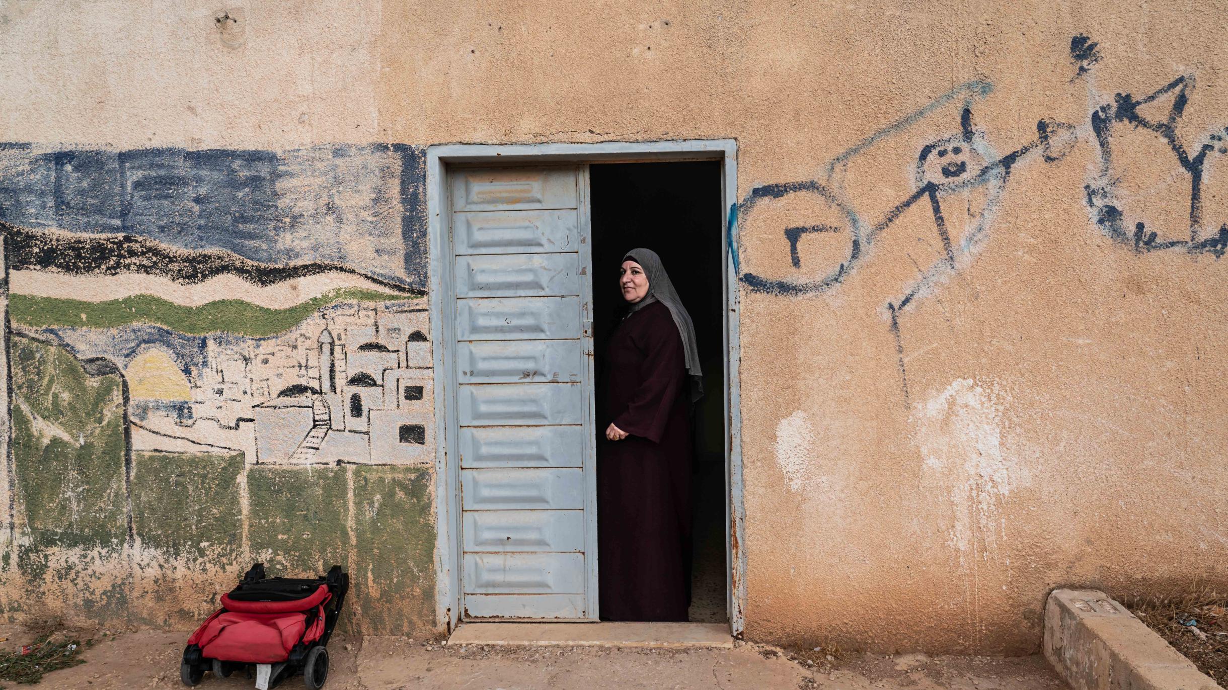 Iman, gekleidet in einem dunklen Gewand, steht in einer hellblau gestrichenen Tür. Die Steinwand um die Tür herum ist mit Schriftzeichen und einem Stadtbild bedeckt.