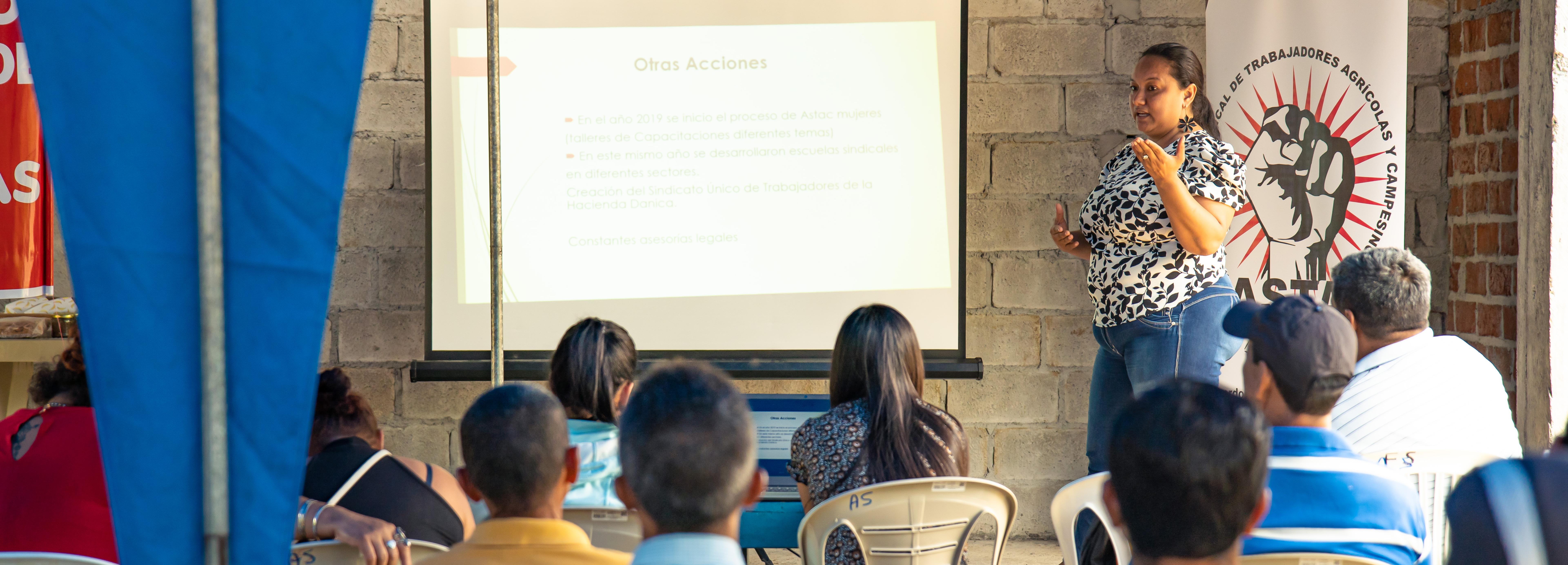 Diana Montoya steht vor einer Grupper Interessierter und erklärt mit Hilfe einer Powerpoint-Präsentation die Arbeit von Gewerkschaften wie ASTAC.