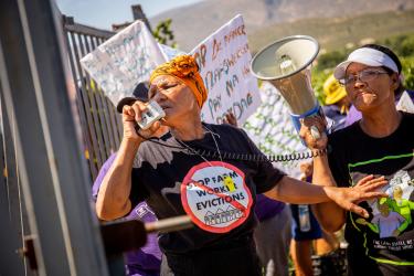Aktivistin Bettie Fortuin leitet einen Streik vor der Matroosberg Farm in De Doorns, Südafrika. Auf der rechten Seite mit Megafon: Magrieta Prins. Die Gruppe fordert eine regelmäßige und pünktliche Bezahlung der Arbeiter*innen auf der Farm. Eine Frau spricht in ein Megafon, die andere hält das Megafon. Aufschrift auf einem T-Shirt: „Stop Farm Worker Evictions“ „(Stoppt die Zwangsräumung von Farmarbeiter*innen“)