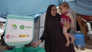 Duaa Abu Sabha benutzt eine Handwaschstation, die Oxfam für Geflüchtete im Gebiet Al-Mawasi im Gouvernement Khan Yunis im Gazastreifen aufgestellt hat. Duaa Abu Sabha wäscht sich die Hände an einer Handwaschstation, die in einem Camp für Geflüchtete steht. Auf dem Arm trägt sie ein Kleinkind.
