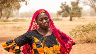 Klimakrise in Burkina Faso: Sanfo Ramata ist Bäuerin und Viehzüchterin. Sie arbeitet für die Confédération Paysanne du Faso, eine Partnerorganisation von Oxfam. Ihr Vieh stirbt wegen des Futtermangels und der Wasserknappheit, die durch die Klimakrise verstärkt werden. Sanfo Ramata ist Bäuerin und Viehzüchterin. Sie arbeitet für die Confédération Paysanne du Faso, eine Partnerorganisation von Oxfam. Die Frau steht in einem bunten Kleid vor einem Haus. Sie sieht nachdenklich aus.