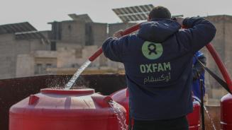 Ein_e Mitarbeiter_in von Oxfam in Syrien befüllt einen Wassertank, damit die Menschen in Notunterkünften Zugang zu sauberem Trinkwasser haben. Ein_e Oxfam-Mitarbeiter_in, mit einer blauen Kaputzenjacke mit grünem Oxfam-Logo bekleidet, befüllt mithilfe eines Schlauches einen roten Wassertank mit sauberem Trinkwasser.