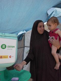 Duaa Abu Sabha benutzt eine Handwaschstation, die Oxfam für Geflüchtete im Gebiet Al-Mawasi im Gouvernement Khan Yunis im Gazastreifen aufgestellt hat. Duaa Abu Sabha wäscht sich die Hände an einer Handwaschstation, die in einem Camp für Geflüchtete steht. Auf dem Arm trägt sie ein Kleinkind.