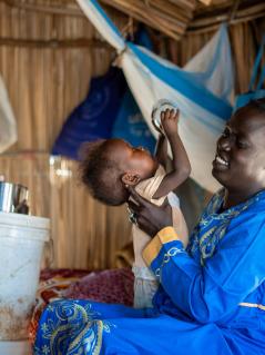 „Als Geflüchtete im Südsudan erhalte ich eine monatliche finanzielle Unterstützung, Hygieneartikel und Seife von Oxfam. Das hilft mir, aber ich würde gerne mehr für die Ernährung meiner Kinder tun.“ Ayul Ajak, Mutter von sechs Kindern und sehbehindert, ist vor dem Krieg im Sudan geflohen. Ayul Ajak sitzt in einer provisorischen Unterkunft und spielt mit einem Kleinkind. Im Hintergrund steht ein Eimer mit der Aufschrift „Oxfam“.