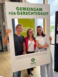 Im Rahmen des Jahresnetzwerktreffens anlässlich des zehnjährigen Bestehens des Netzwerks "Unternehmer*innen für Unternehmer*innen" trafen sich Unternehmer Jens Seidel, Oxfam-Mitarbeiterin Annika Wild und Unternehmerin Nell Seidel (von links) für Gespräche und Austausch. Drei Personen halten einen Rahmen mit der Aufschrift „Gemeinsam für Gerechtigkeit“.